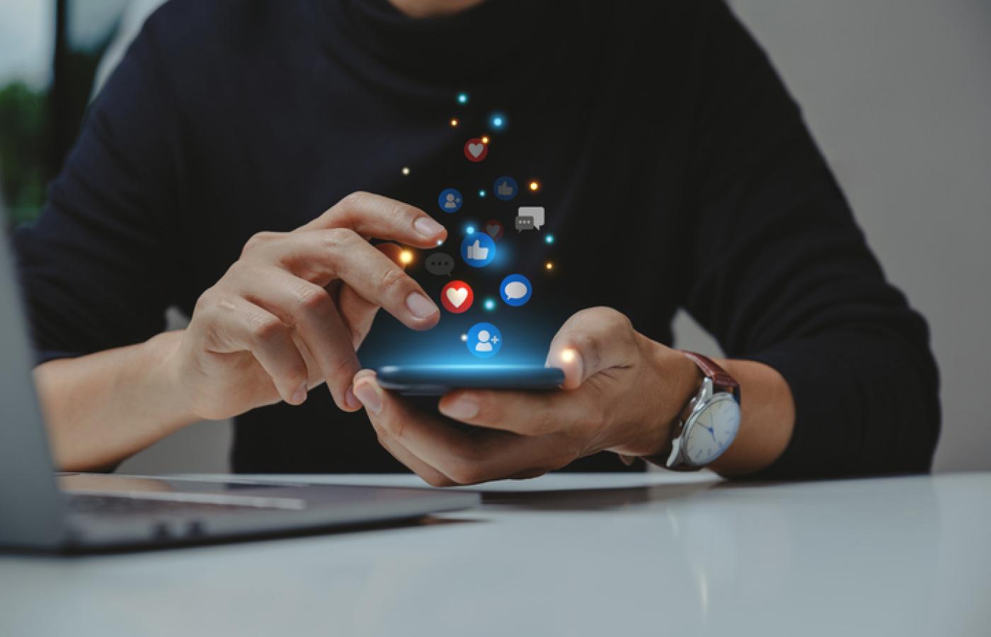 Hands tapping on a mobile phone with social media icons glowing above the phone.